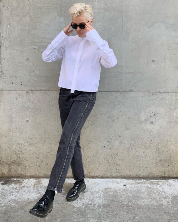 Brittenelle kicking one leg out and adjusting her sunglasses with her hands in front of a cement wall.